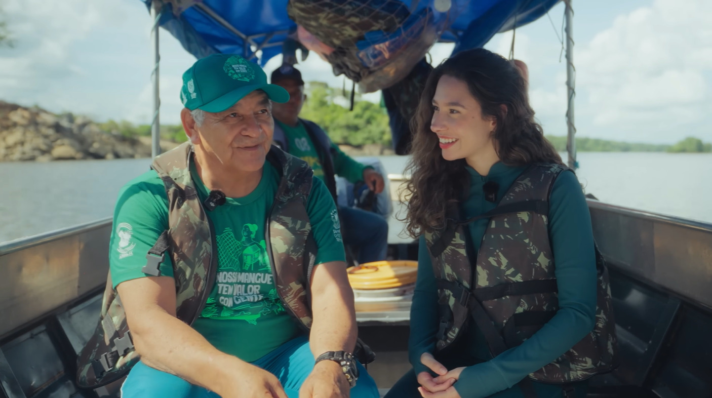 Mulher e homem com colete em um barco. Ela olha para ele enquanto ele fala.