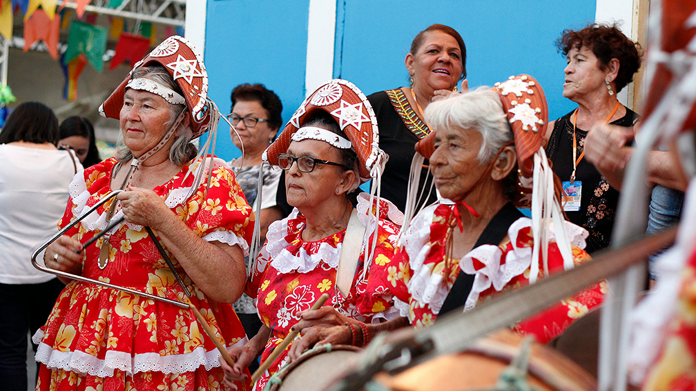 Três mulheres idosas celebram São João com roupas típicas e tocando instrumentos musicais.
